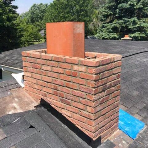Brick chimney on a shingled roof with a rusty metal cap and trees in the background