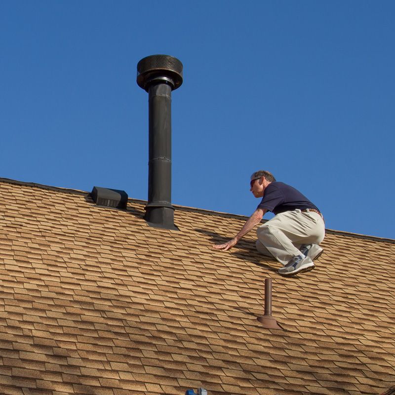 Worker crouching on a shingled roof beside a metal vent pipe under a blue sky