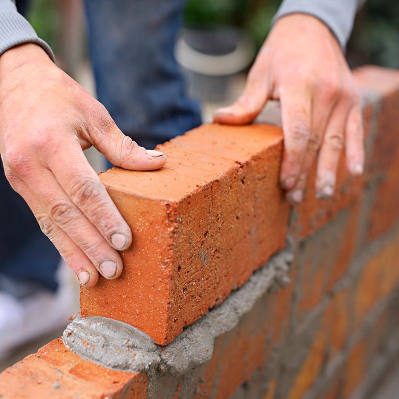 Hands placing a red brick onto a wet mortar chimney wall during construction