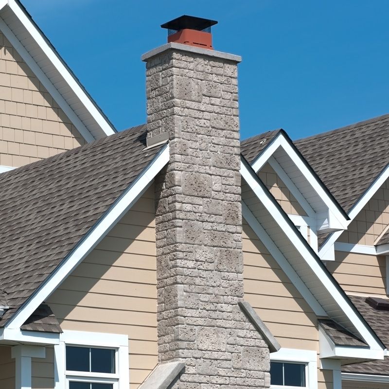 Stone chimney on a beige house with dark shingle roofs and white-trimmed gables under a blue sky