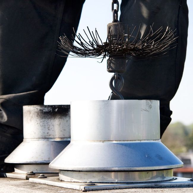 Two metal chimney caps being cleaned with a wire brush, seen between a worker’s legs.