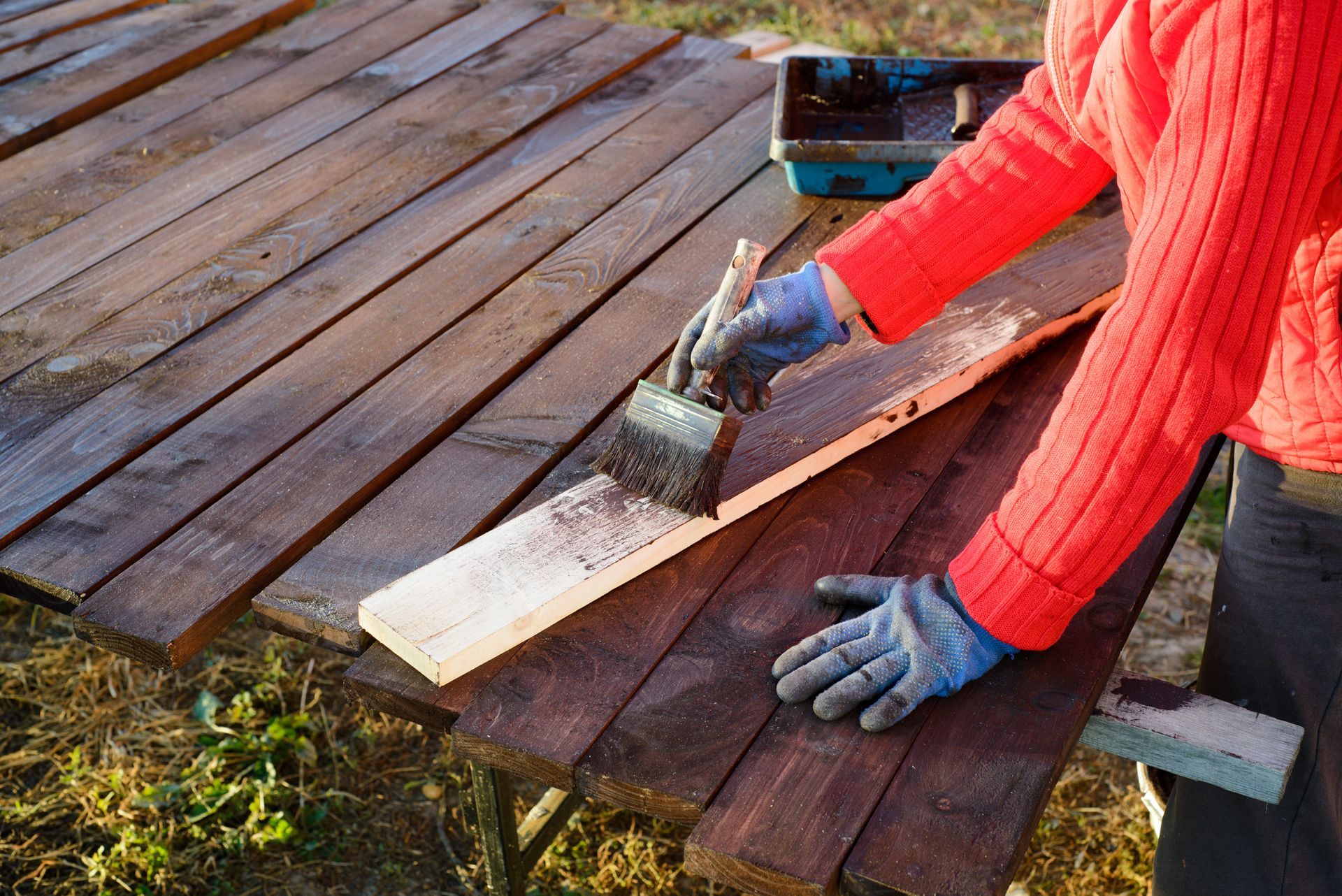 Person in red sweater refinishing a wooden deck with a paintbrush and gloves