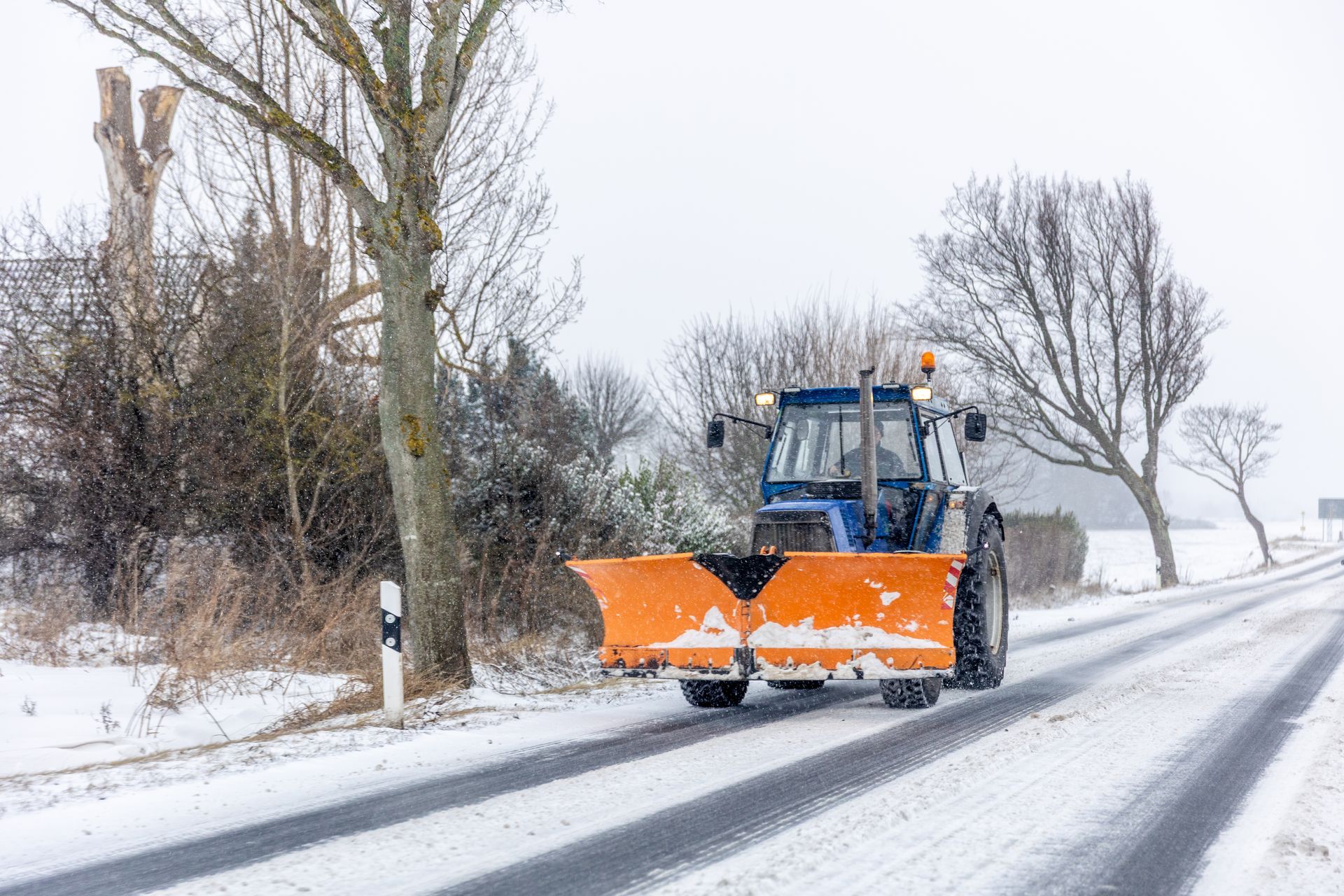 Orange snowplow clearing a snowy road lined with bare trees