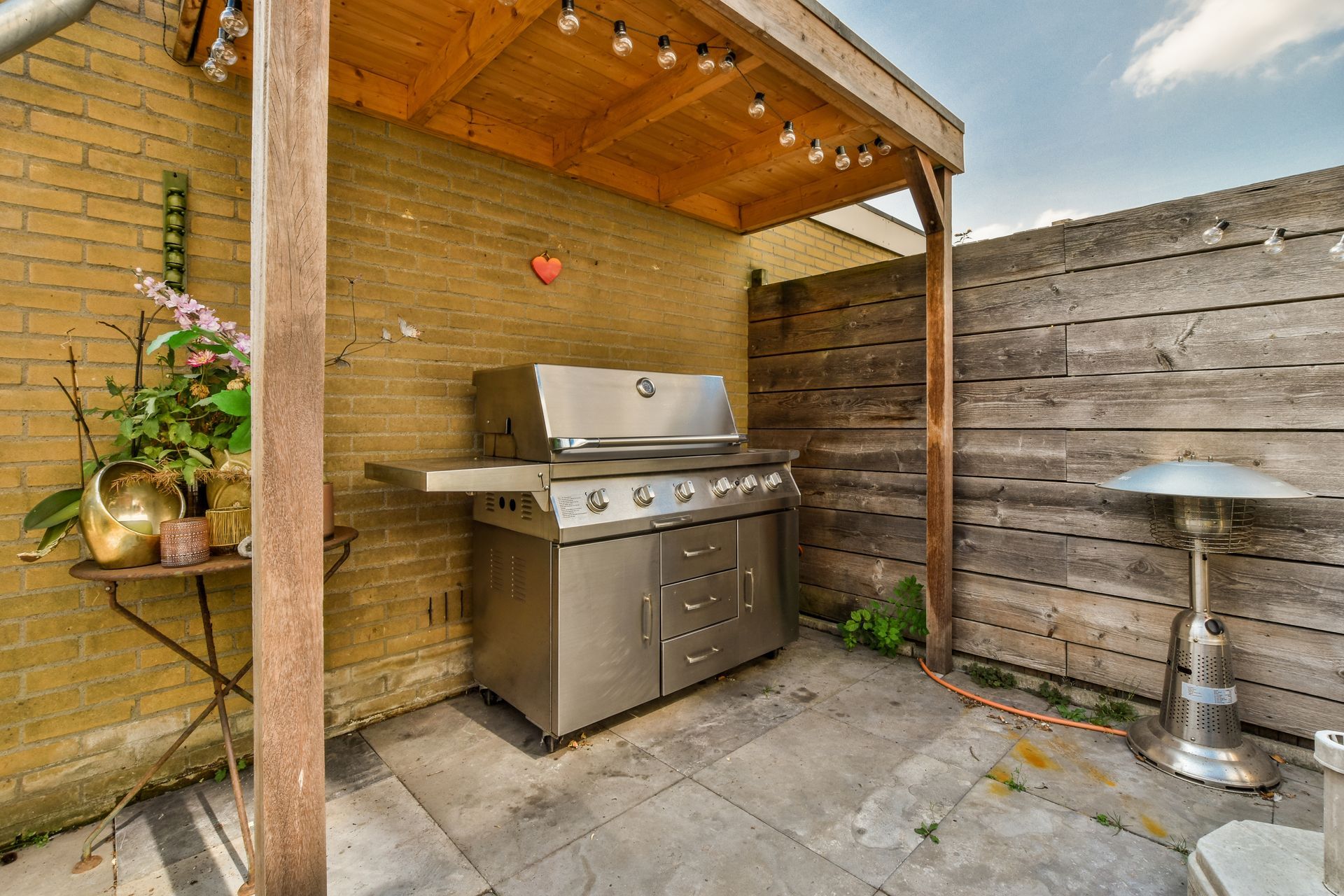 Outdoor patio with stainless steel grill under a wooden pergola, stone pavers, and a metal stool.