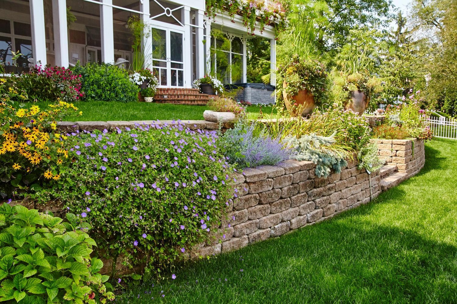 A stone retaining wall built into a grassy hill, fronted by colorful flowers, with a white house in the background.