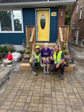 Three workers in high-visibility vests sit on front steps of a house with a yellow door and brick walkway.