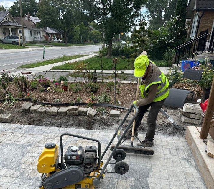 Worker in neon safety gear using a compactor on a stone patio beside a garden and residential street.