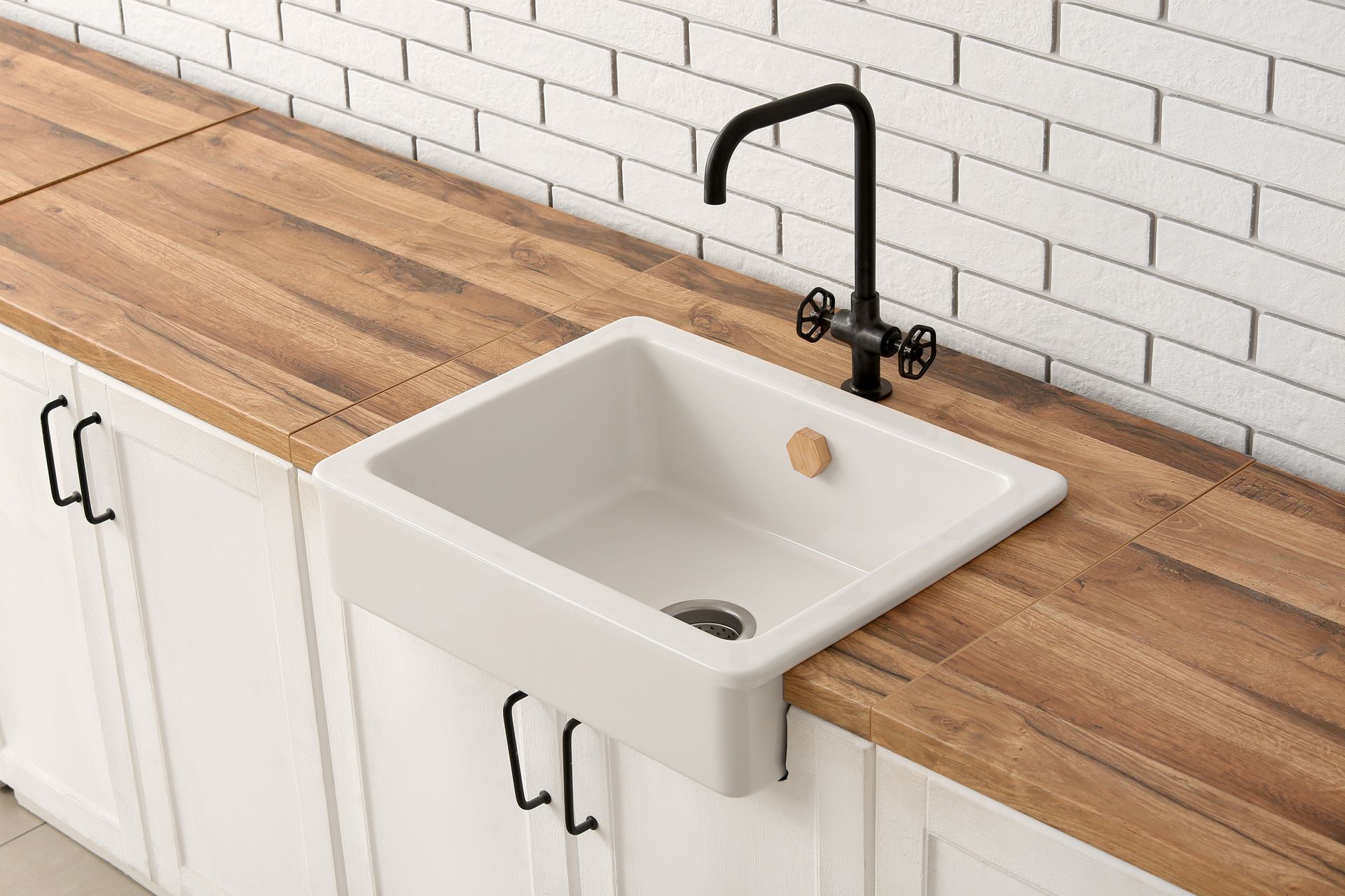 White farmhouse sink with black faucet set in a wooden kitchen countertop against a white tile wall