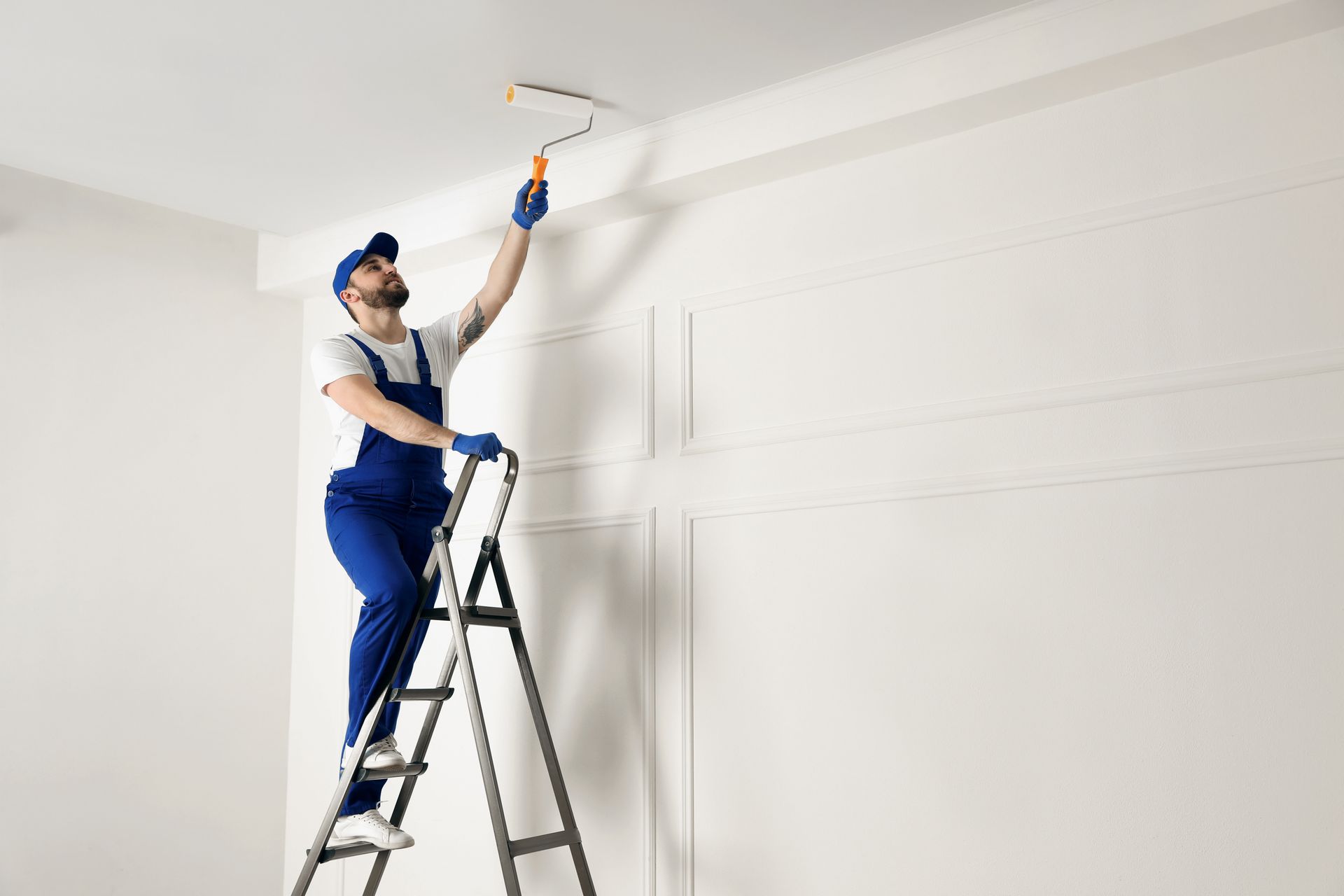Painter on a ladder rolling white paint onto a high wall in a bright room