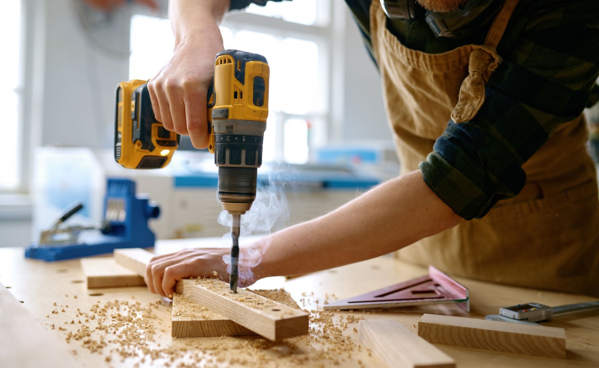 Hands using a yellow drill to bore into a wooden plank on a workbench with sawdust