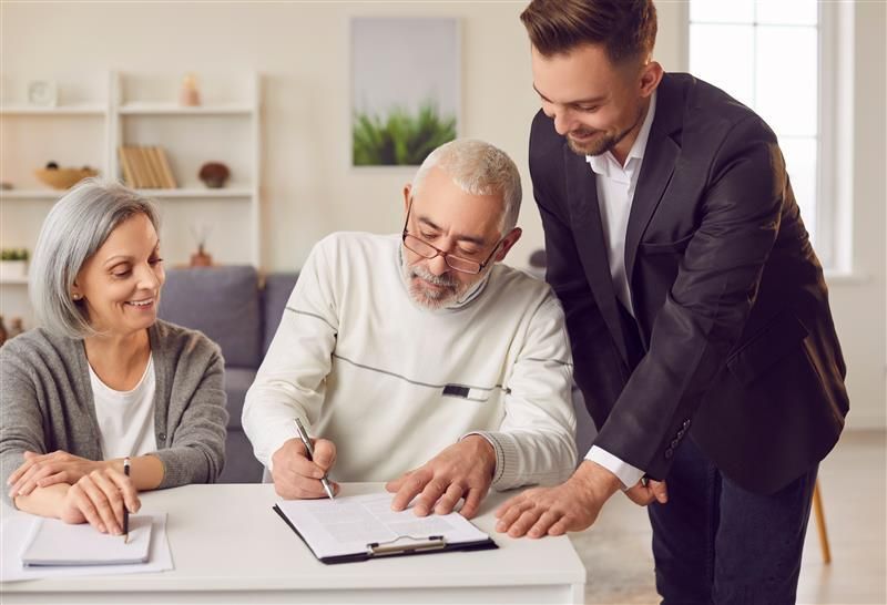 An older couple signing a document at a table with a younger man in a suit assisting them in a home setting.