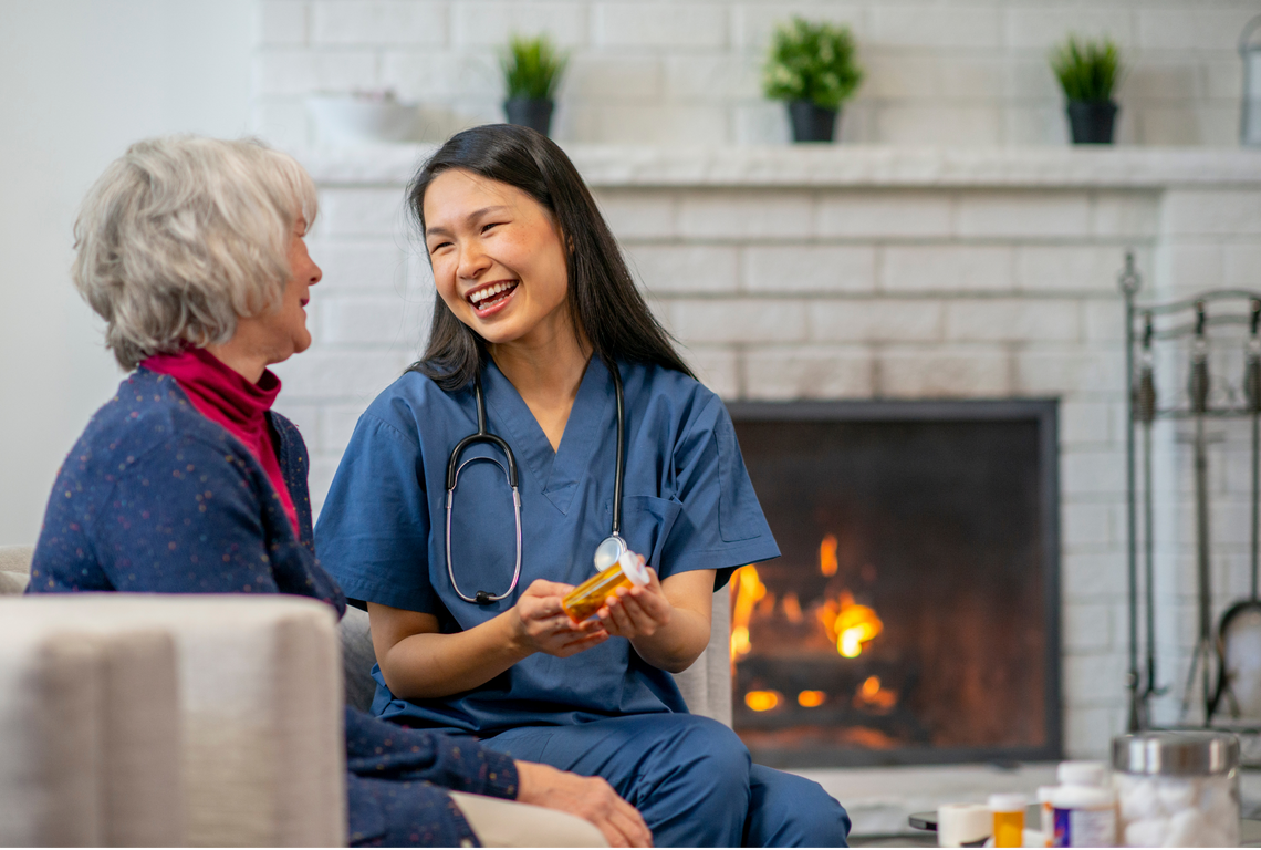 A nurse is talking to an elderly woman in front of a fireplace.