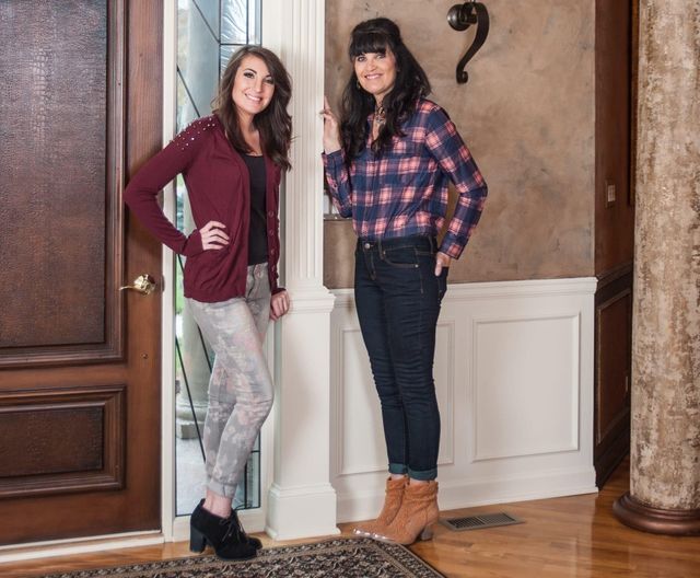 Two women pose indoors, near a doorway. One wears a burgundy sweater and printed pants, the other a plaid shirt and jeans.