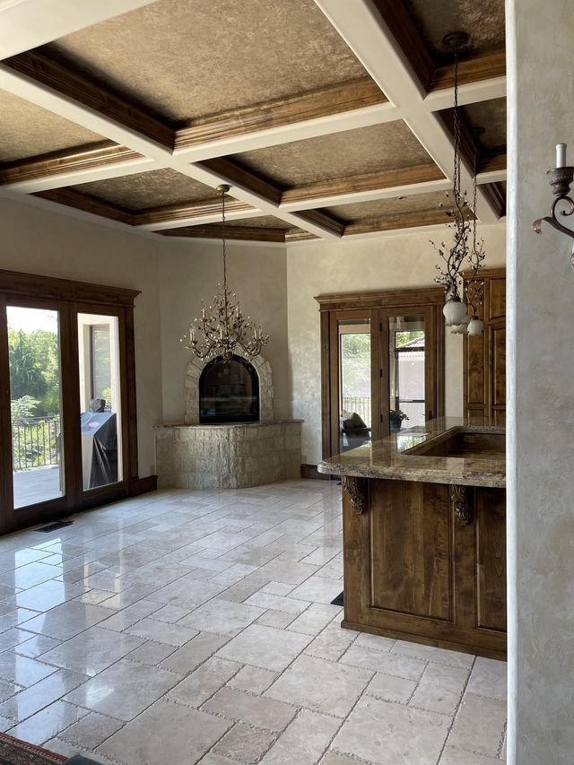 Beamed ceiling with chandelier, fireplace, and kitchen island in a spacious, light-filled interior.