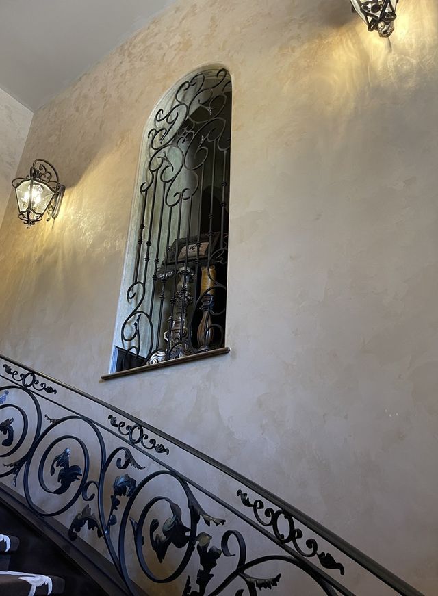 Ornate wrought-iron staircase with matching window bars, sconces on a textured, light-colored wall.
