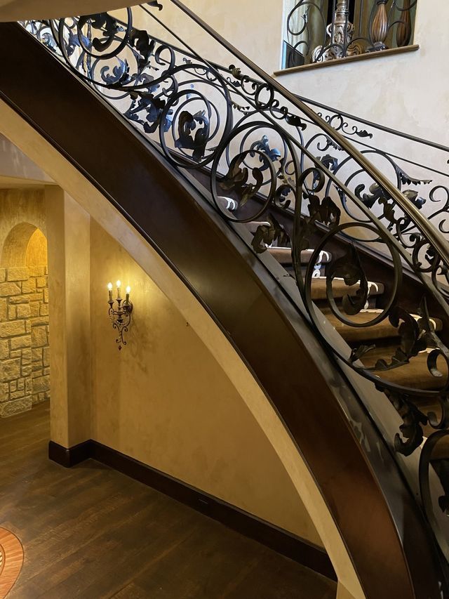 Dark wooden staircase with ornate wrought iron railing. Light tan walls, stone archway, sconce.