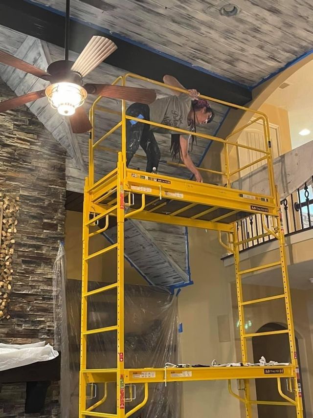 Woman on a yellow scaffold painting a ceiling, wearing casual clothes. Interior shot with stone fireplace and ceiling fan.