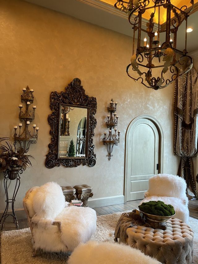 Luxurious living room: cream-colored walls, ornate mirror, fluffy white chairs, chandelier, and a decorative ottoman.
