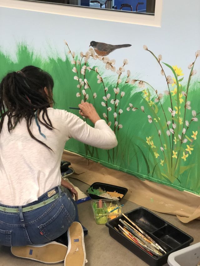 Woman painting a mural of a bird, grass, and flowers on a wall with green, white, and yellow colors.