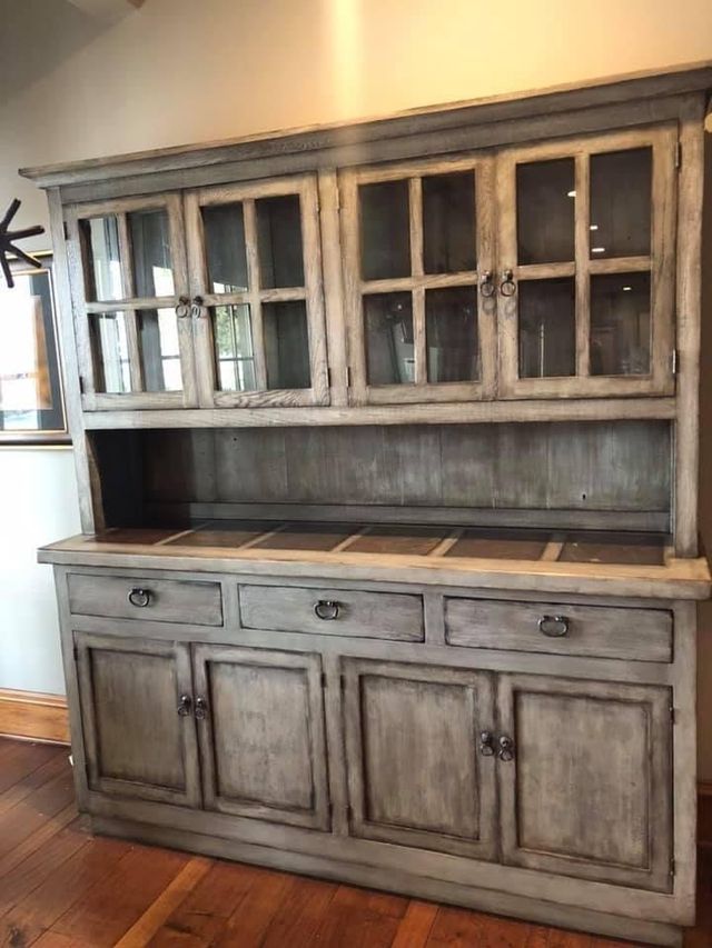 Gray rustic wooden hutch with glass-door upper cabinet, drawers and cabinet doors below. Set on hardwood floor.