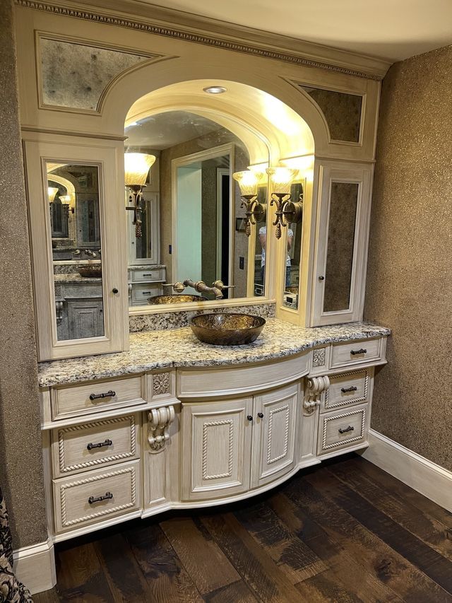 Custom white vanity with granite countertop, curved front, arched mirror, sconces, and cabinets in a bathroom.