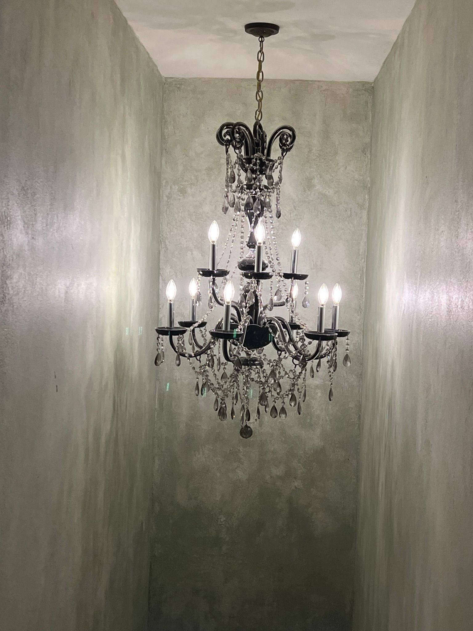 Black wrought-iron chandelier with lit candles hanging in a narrow textured gray hallway