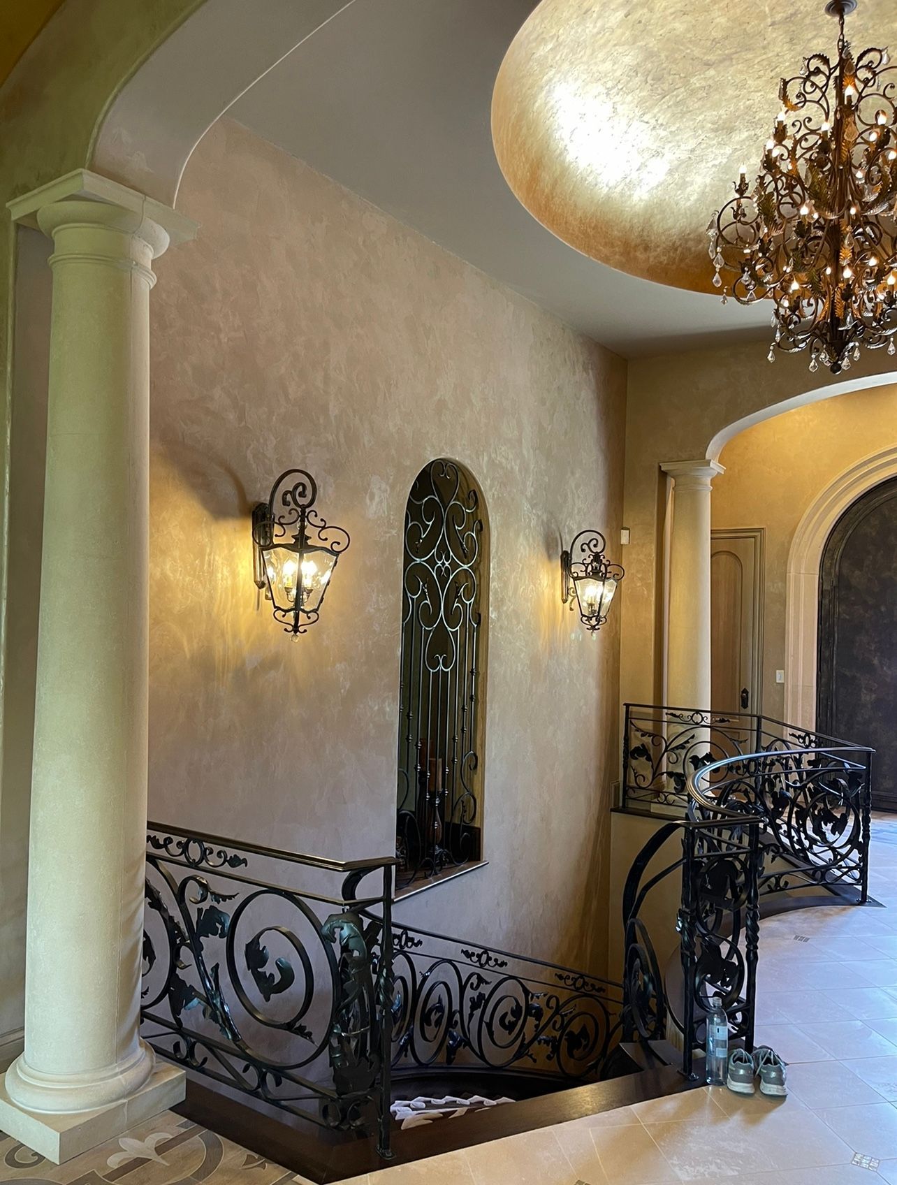 Elegant stucco staircase landing with wrought-iron railing, wall sconces, and chandelier under an arched ceiling