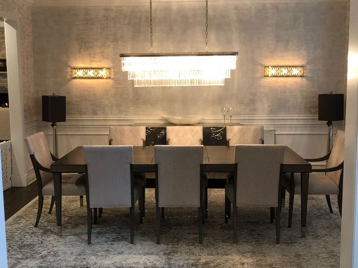 Elegant dining room with long table, upholstered chairs, wall sconces, and a large chandelier over a gray patterned rug
