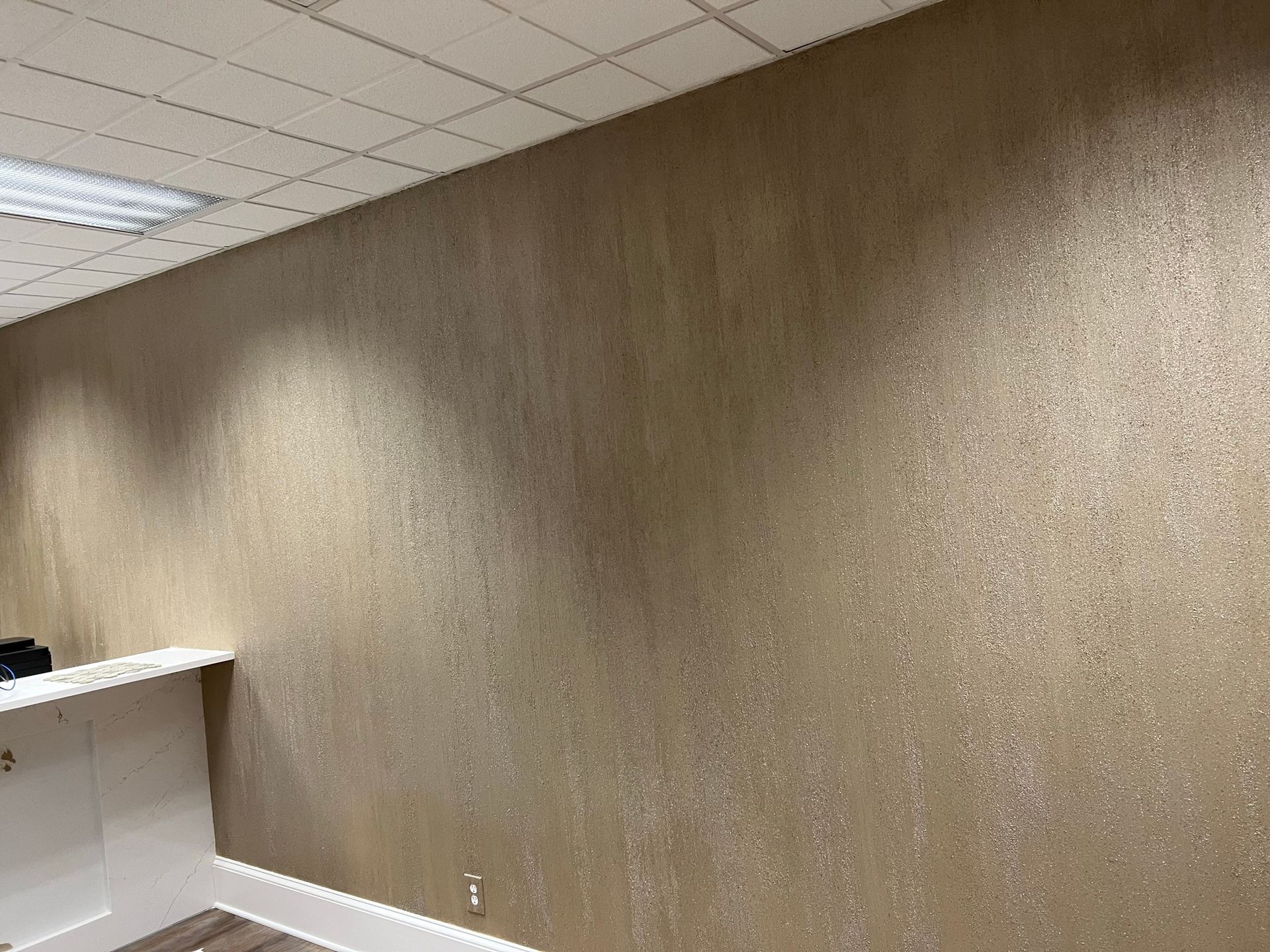 Empty office corner with beige textured wall, white trim, and drop ceiling tiles