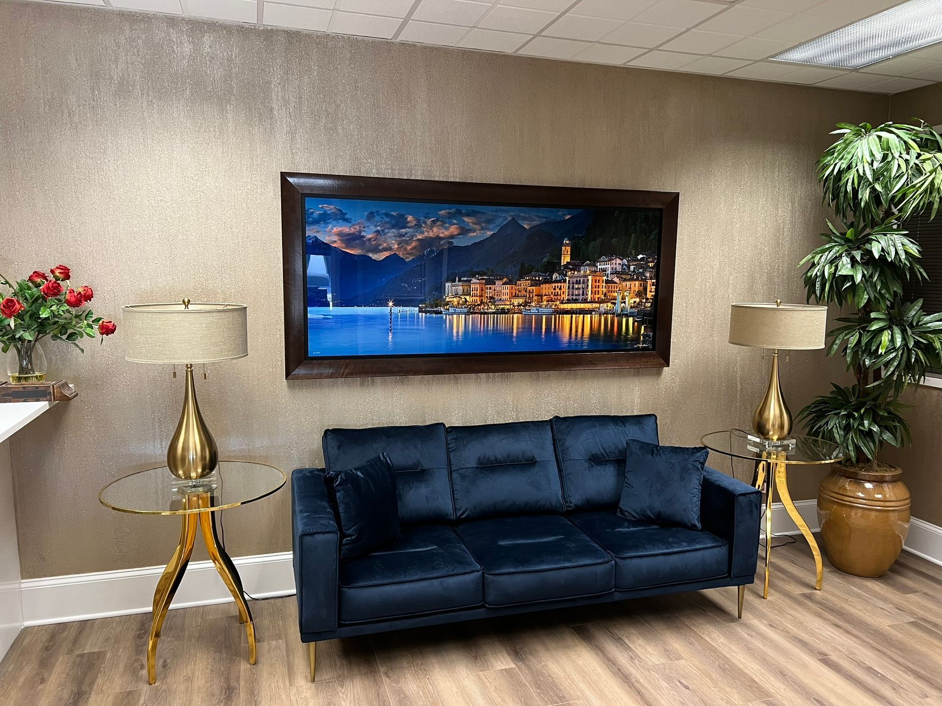 Modern office lounge with navy sofa, framed cityscape, lamps, and potted plants.