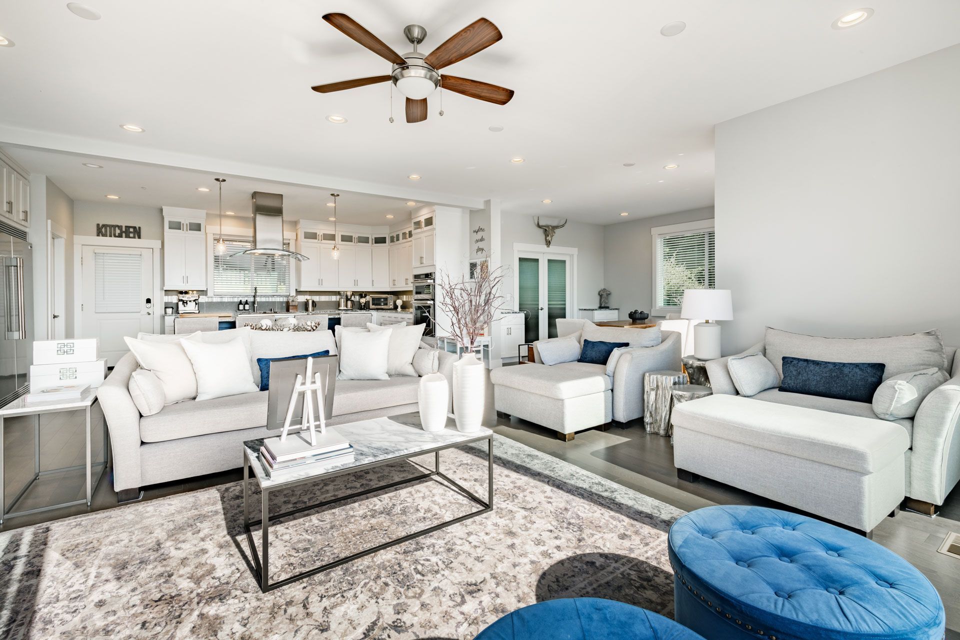Spacious living room with open kitchen. Gray sofas, rug, and ottomans. White walls and ceiling fan.