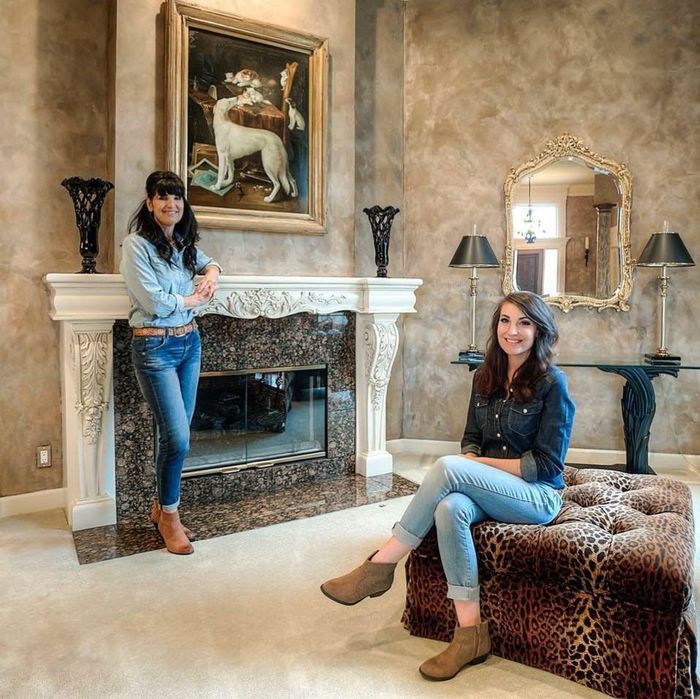 Two women pose in a living room with a fireplace, painting, mirror, and leopard print ottoman.