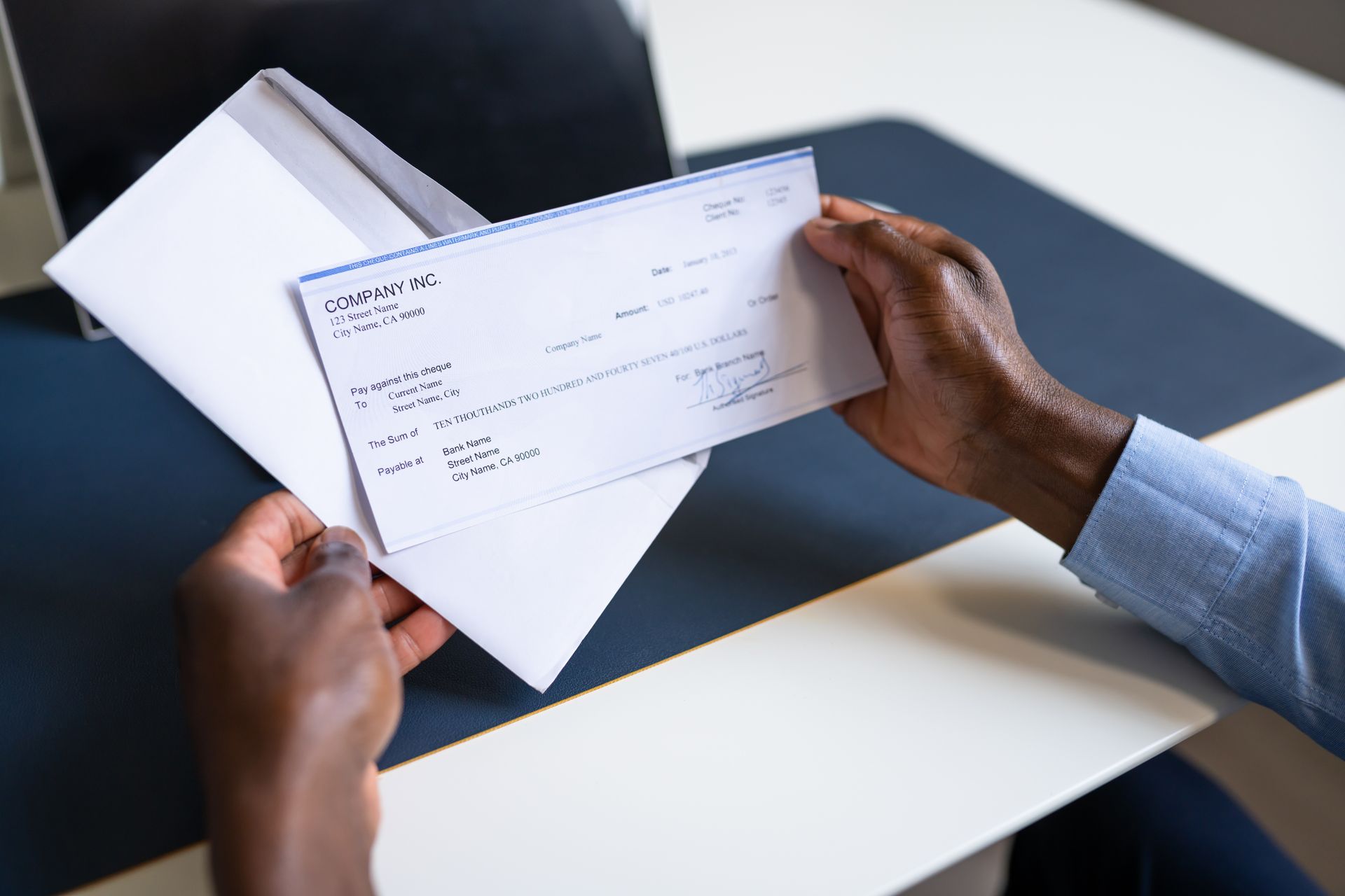 Hands holding a white check over a desk with a laptop in the background