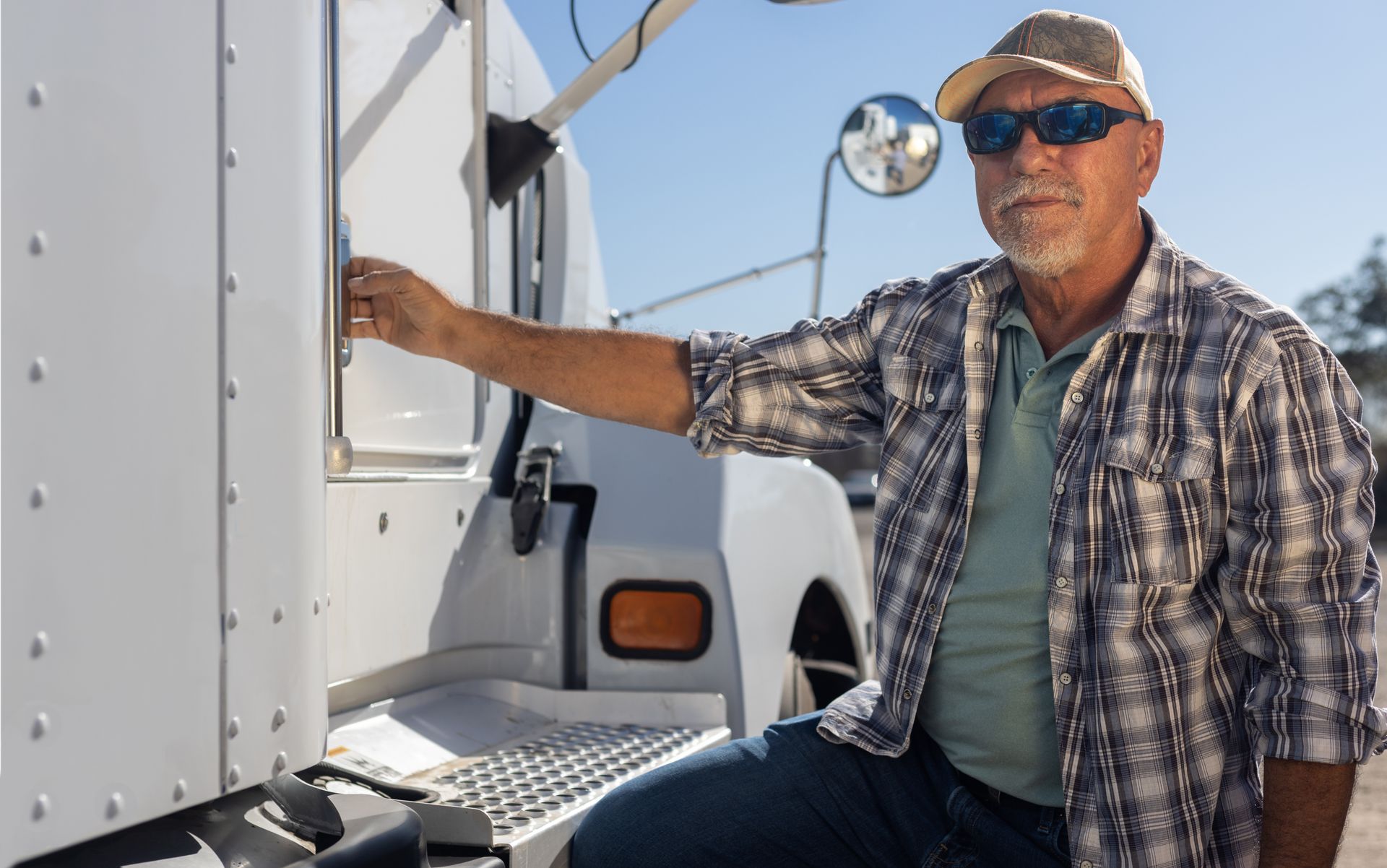 Man in plaid shirt and cap standing by white truck, holding the side door open under bright sunlight.
