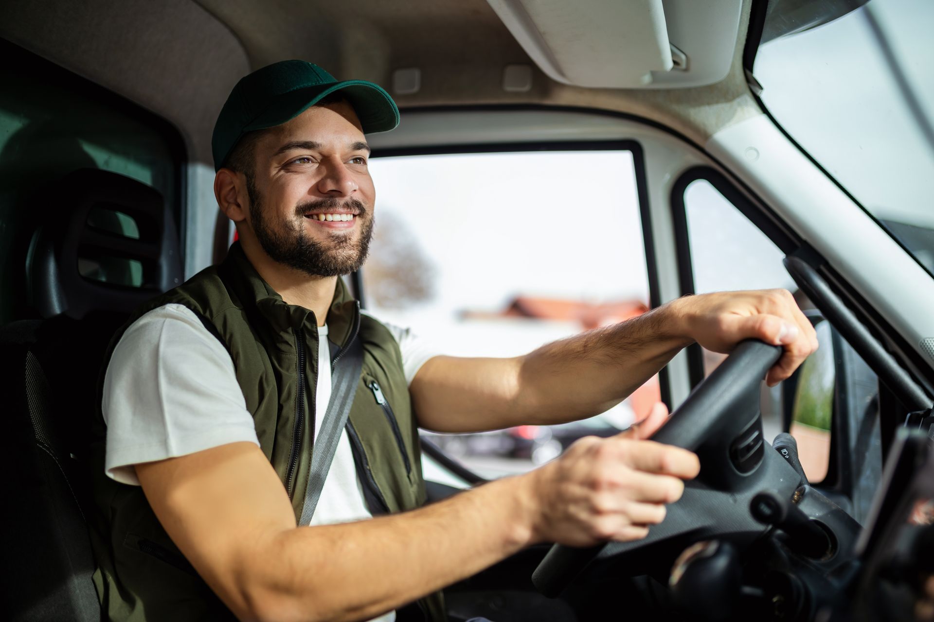 Smiling delivery driver in a van, wearing a cap and seatbelt, driving with one hand on the wheel.