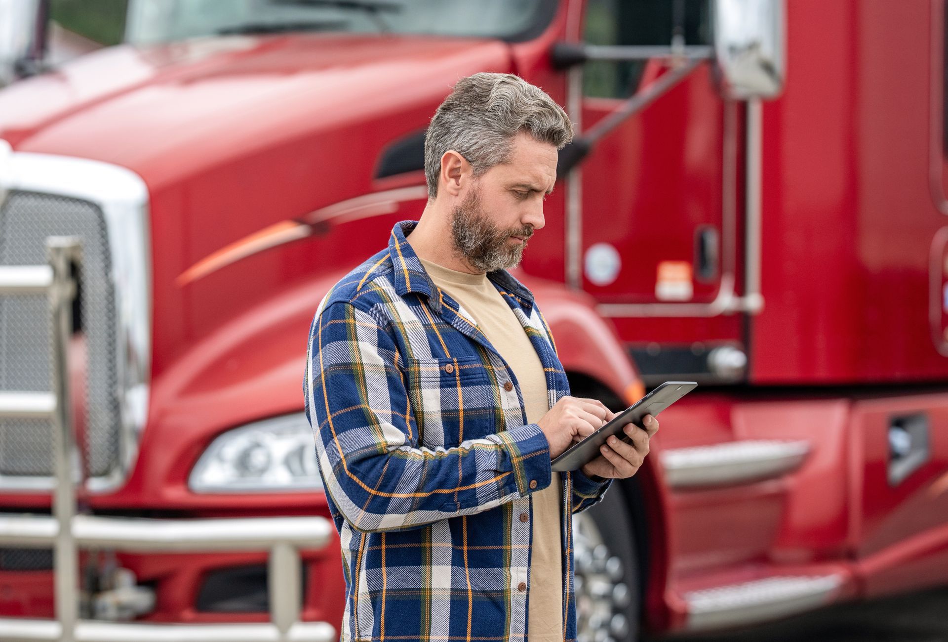 Man in plaid shirt using a tablet beside a red semi-truck