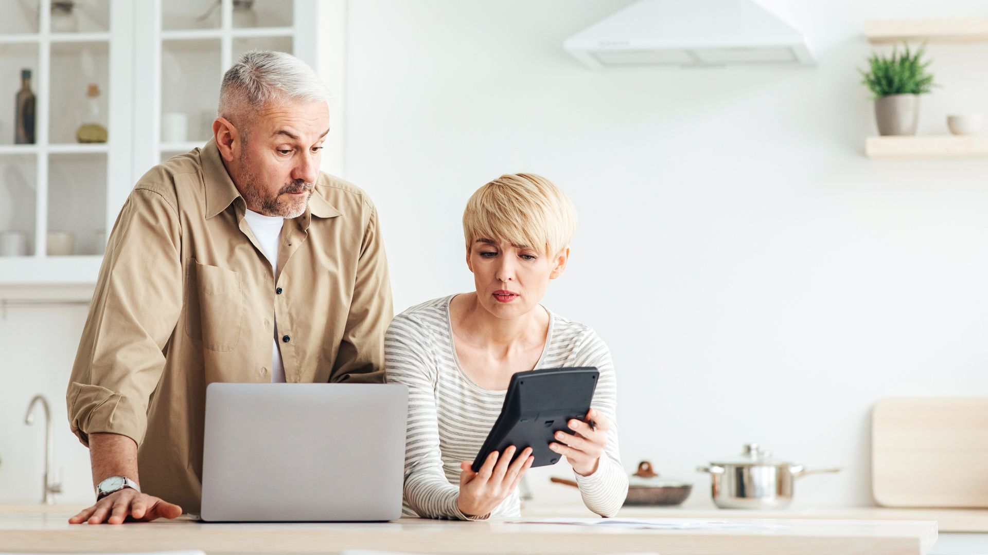 Two people in a bright kitchen looking at a laptop and tablet on the counter.