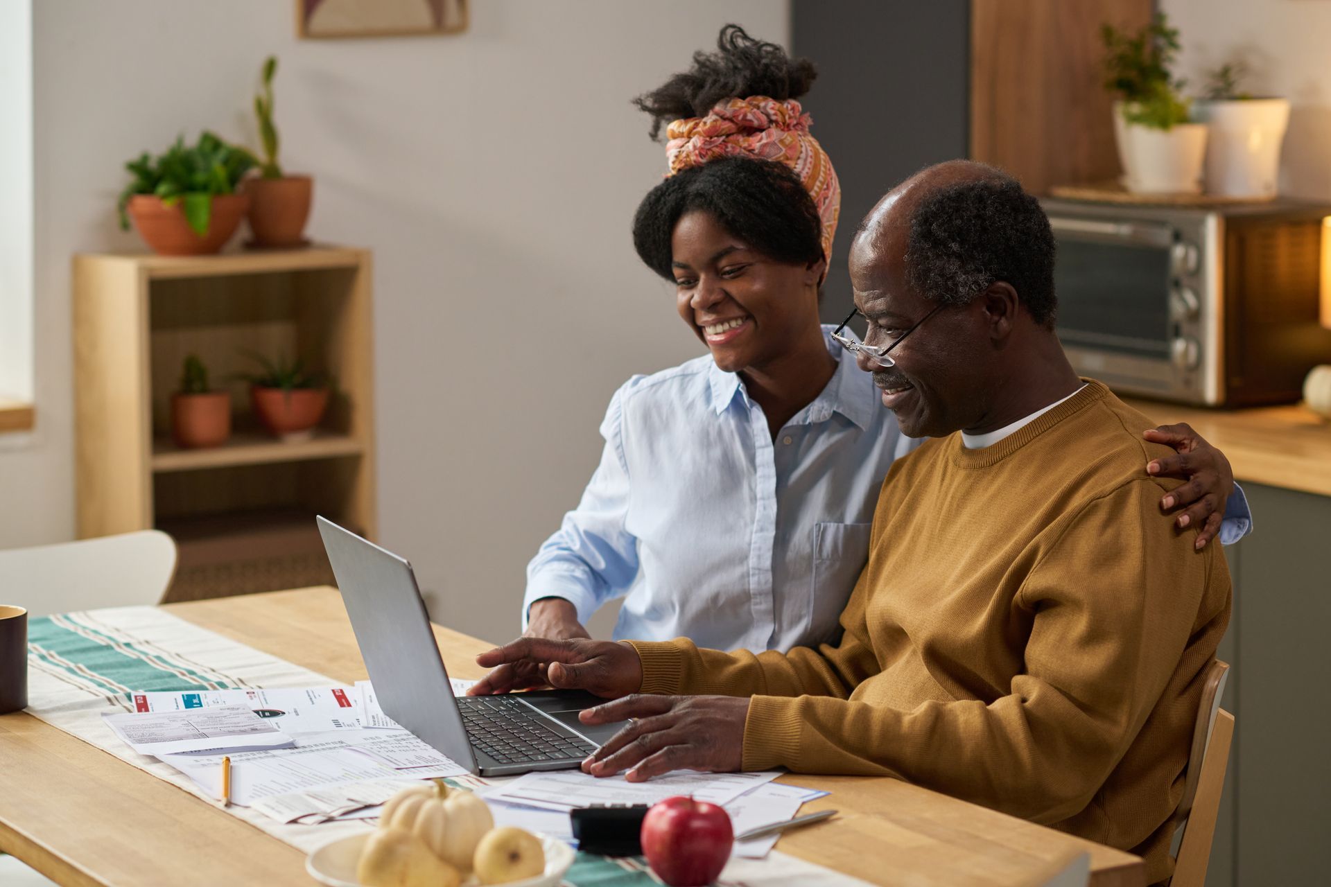 Two people smiling at a laptop on a table in a cozy kitchen, with notebooks and fruit nearby