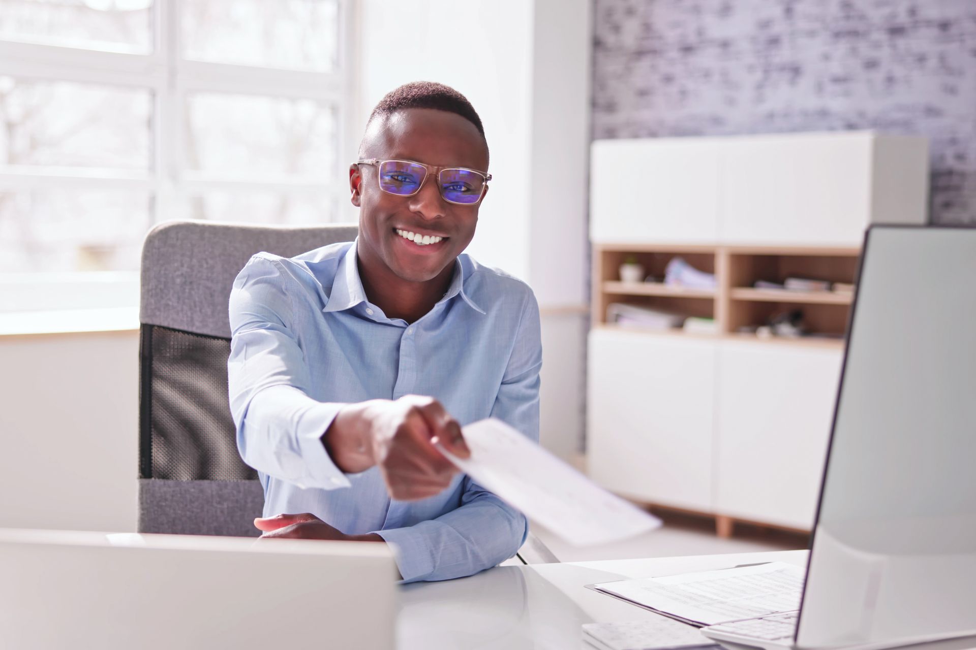 Smiling person in blue shirt handing a paper across a desk in a bright office