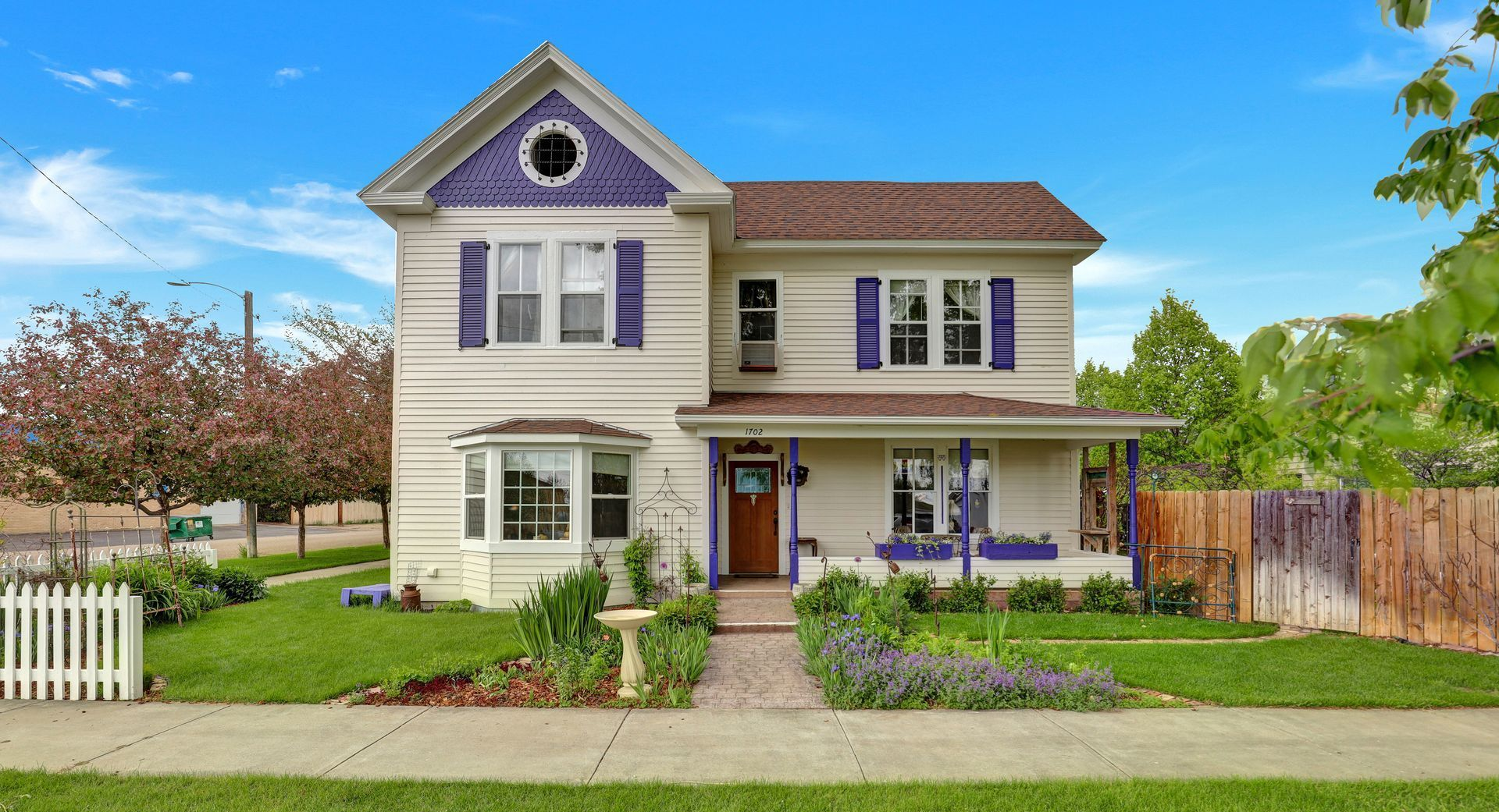 A two-story, white farmhouse-style home with purple trim, a front porch, and a landscaped yard, set under a blue sky.