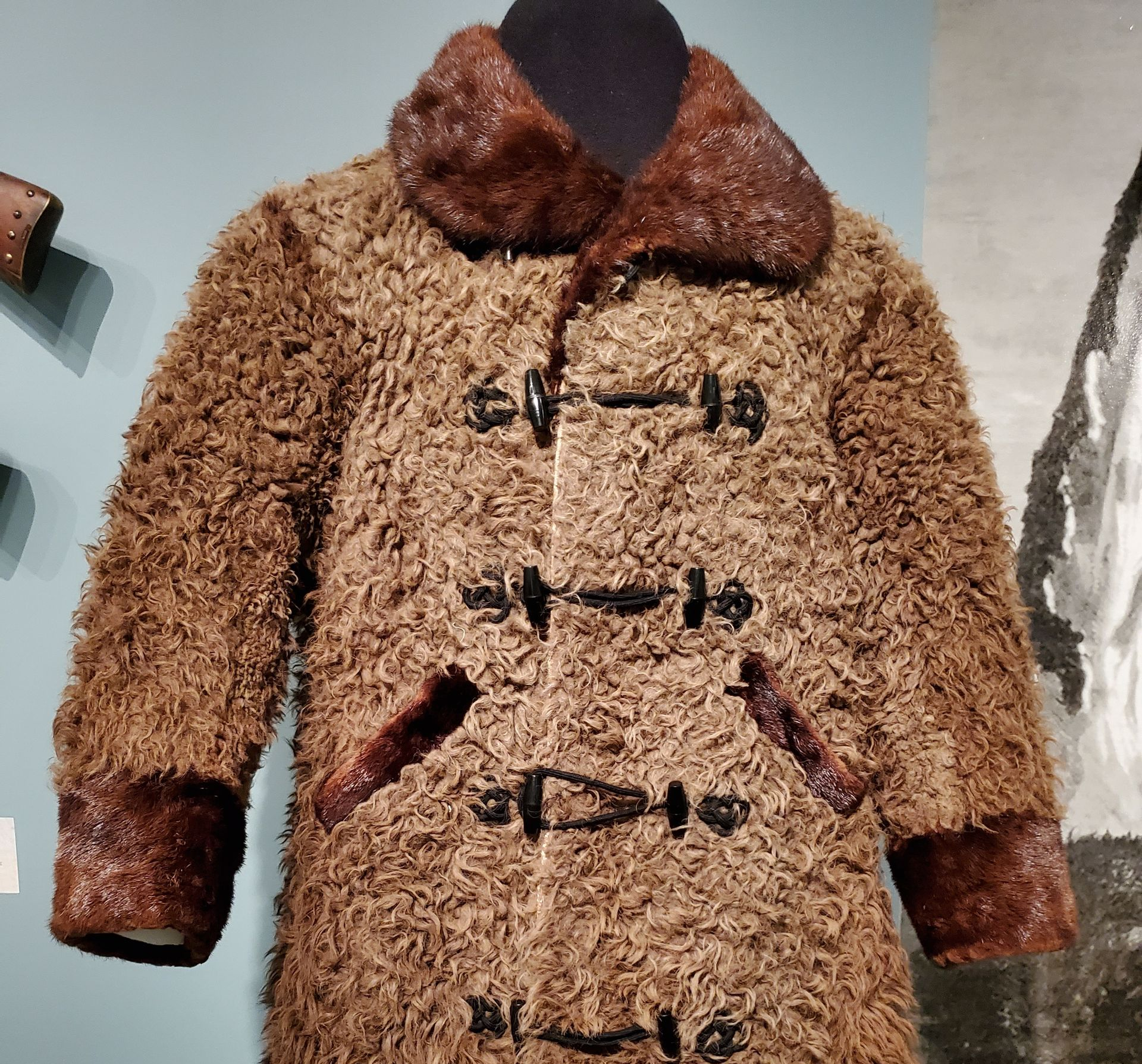 A long, brown shaggy fur coat with dark leather toggle fastenings, a wide collar, and matching cuffs on a display mannequin.