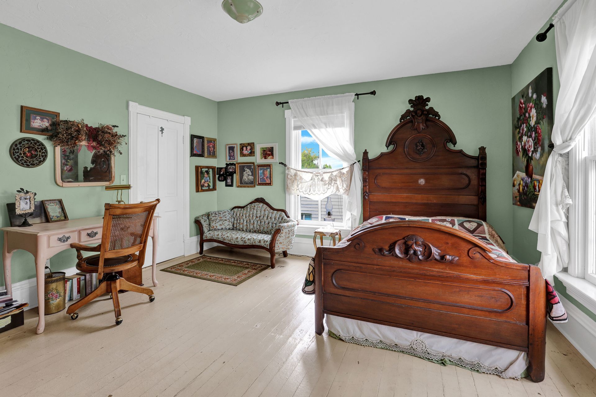 A bedroom with sage green walls, a large ornate wooden bed, a writing desk, and a small patterned settee.