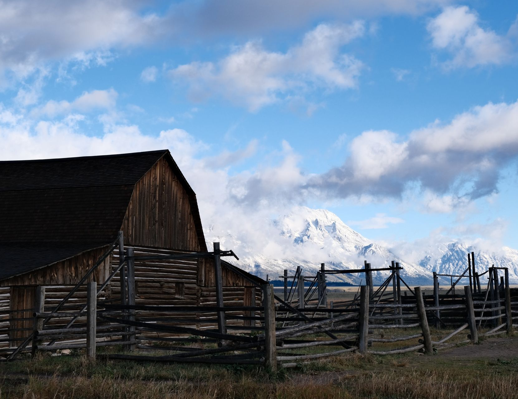 A rustic wooden barn and fence in a field with snow-capped mountains under a blue, cloudy sky.