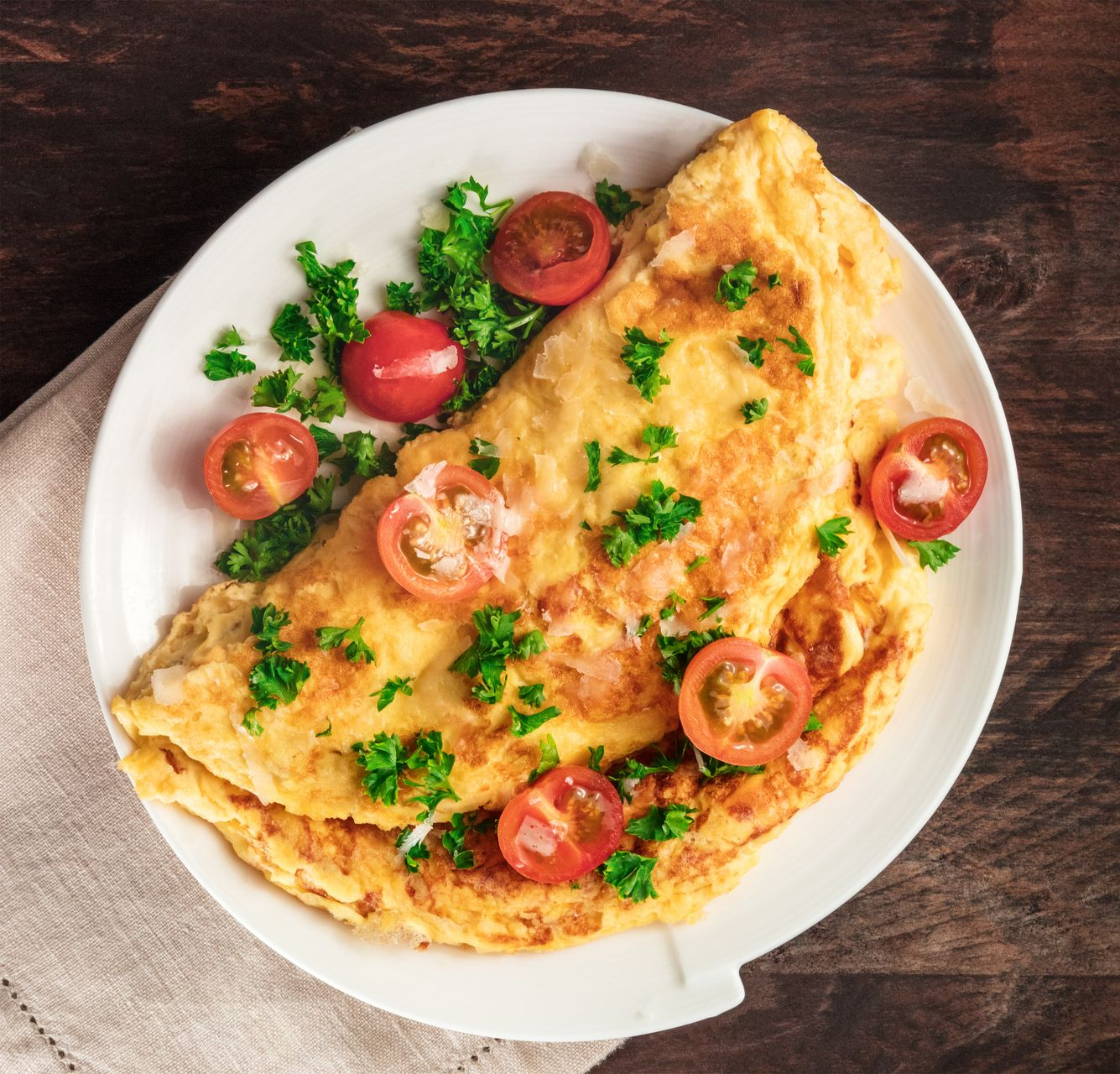 A folded omelet garnished with fresh parsley and sliced cherry tomatoes on a white plate over a dark wooden surface.