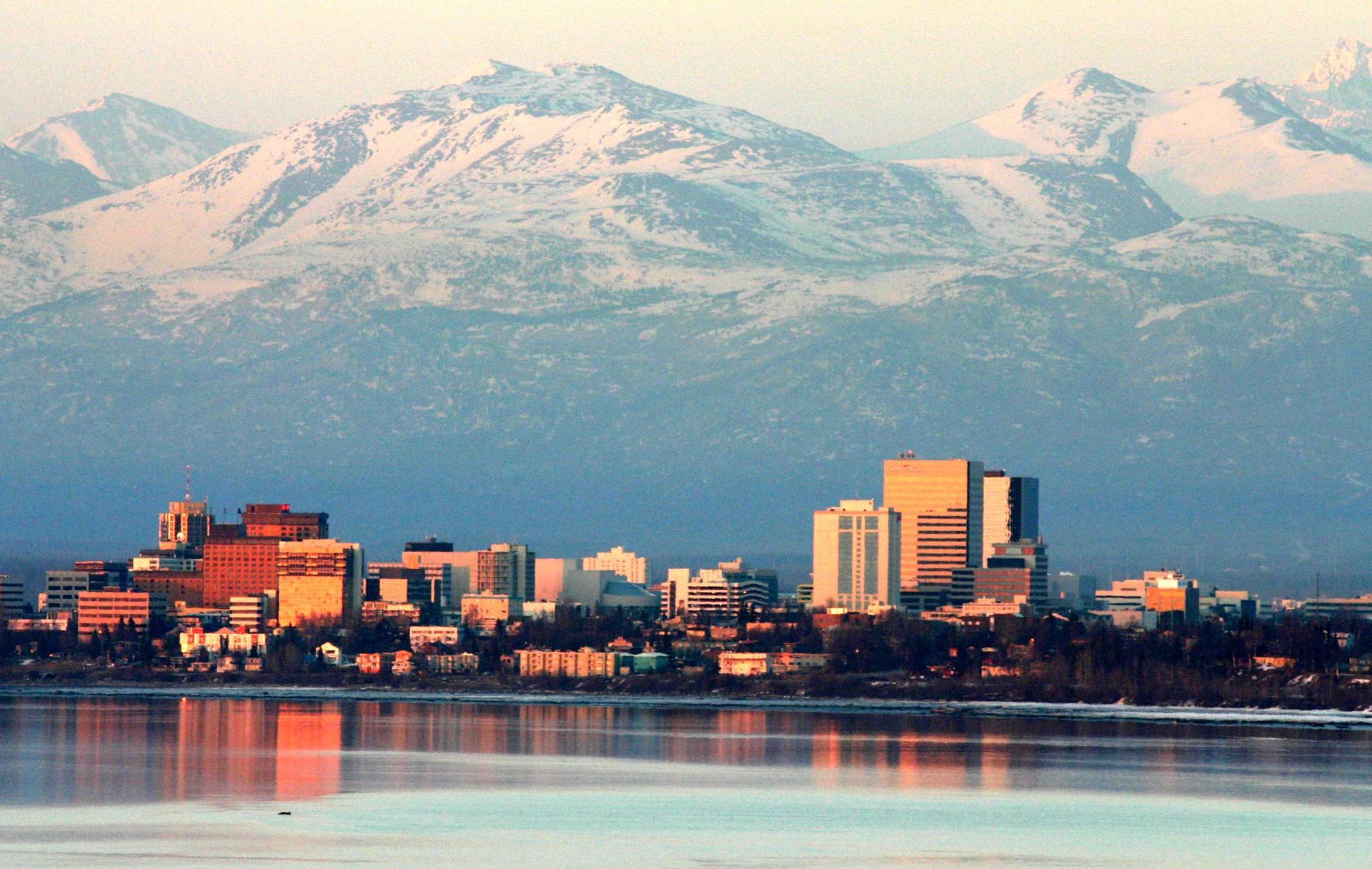 Anchorage skyline with snowy mountains in the background, reflected in calm water.