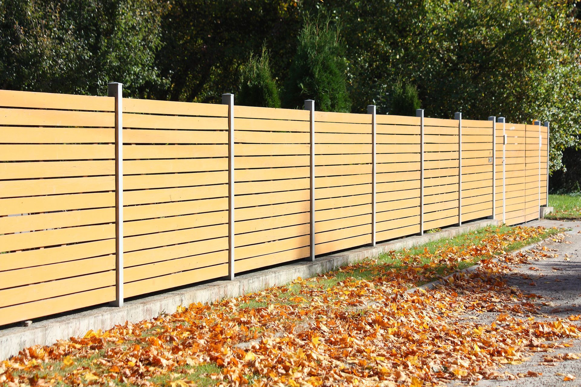 Horizontal slatted wooden fence with metal posts in front of trees and fallen leaves.