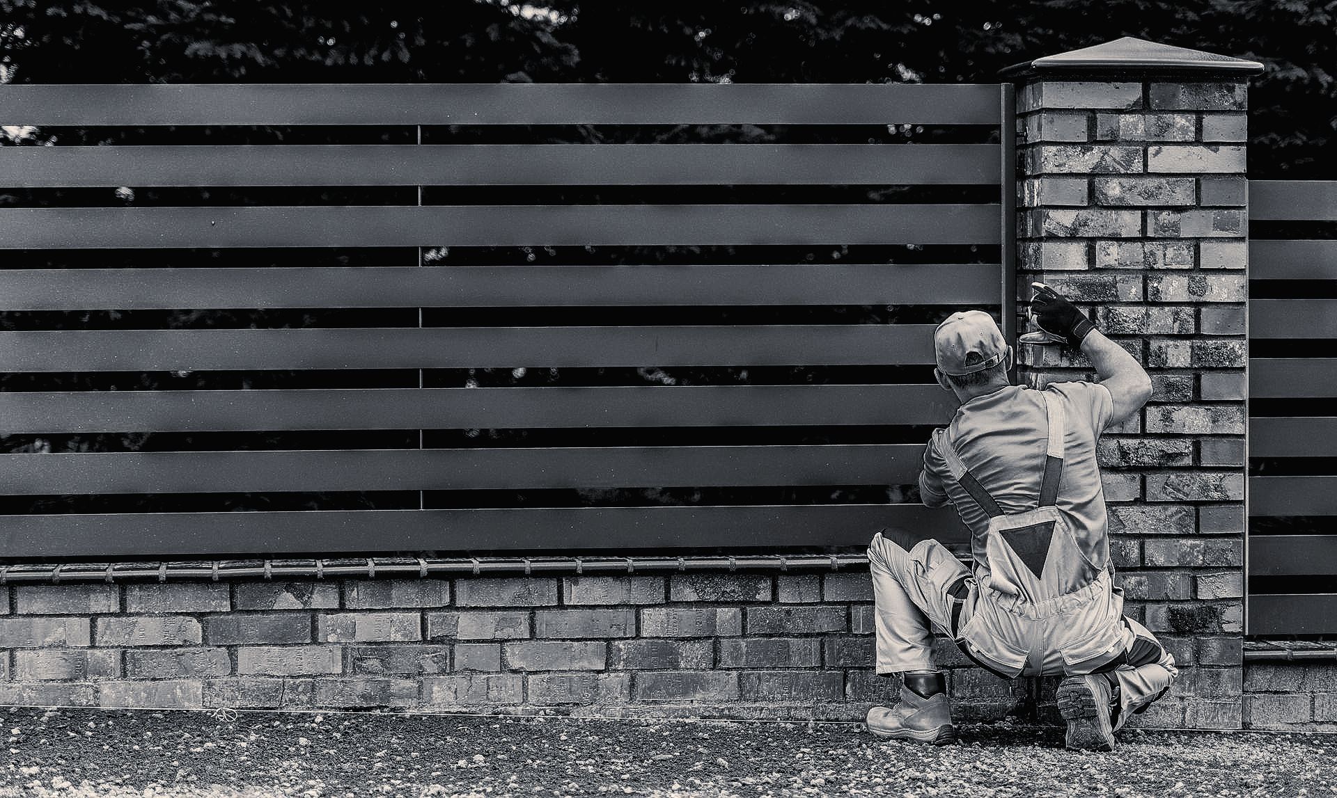 Person crouches, working on a brick fence post next to a dark horizontal slat fence.