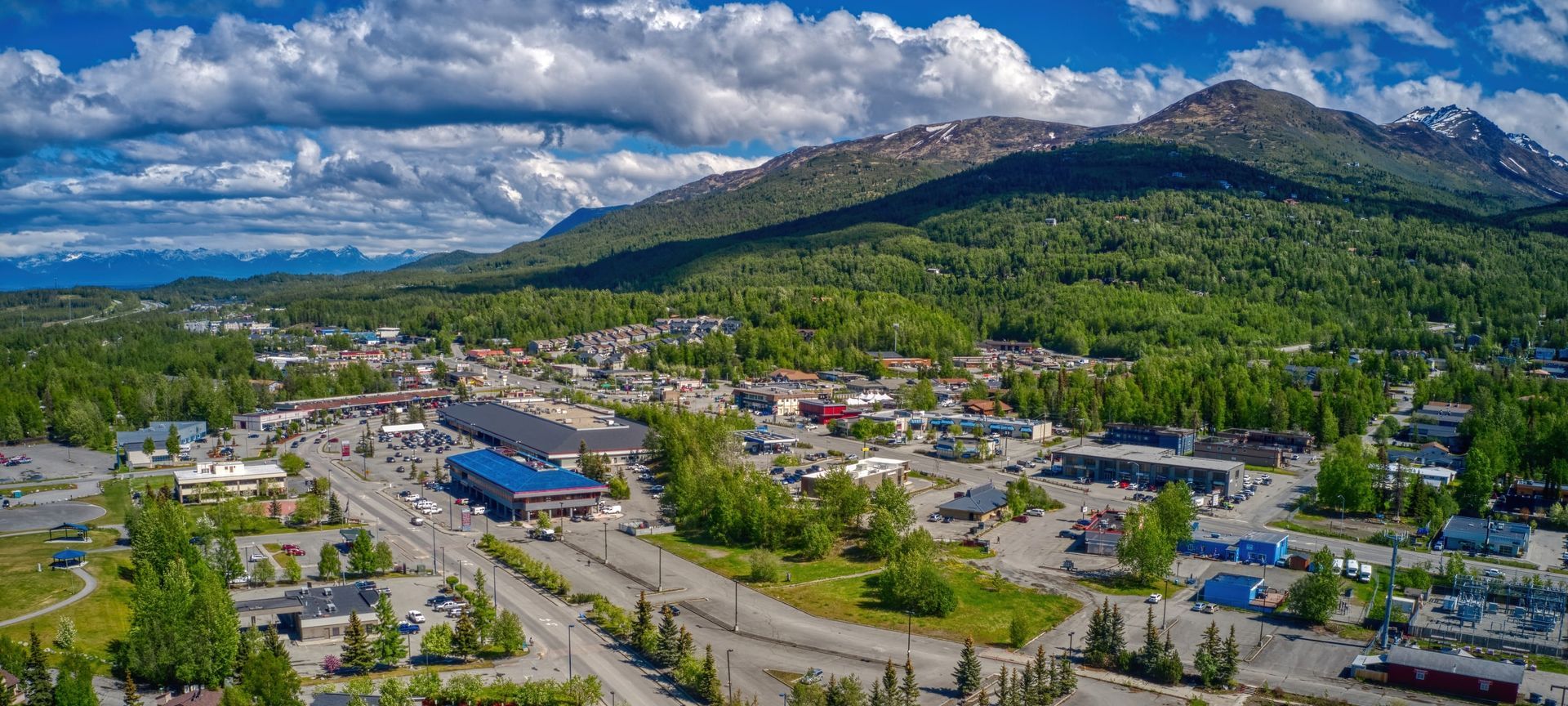 Aerial view of a Eagle River, AK. Lush green trees and mountains under a cloudy blue sky.