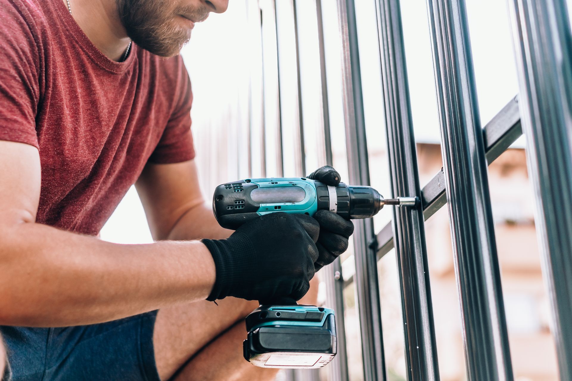 Man in red shirt using a teal drill on a black fence. He wears black gloves and works outside.