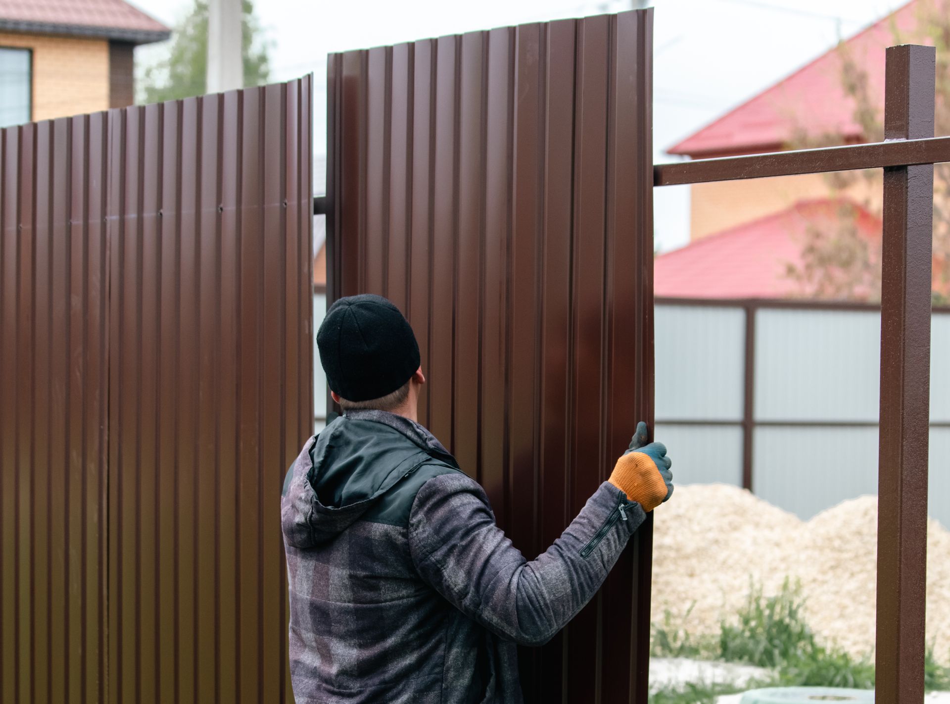 Man installing a brown corrugated metal gate outdoors, wearing a black cap and work gloves.